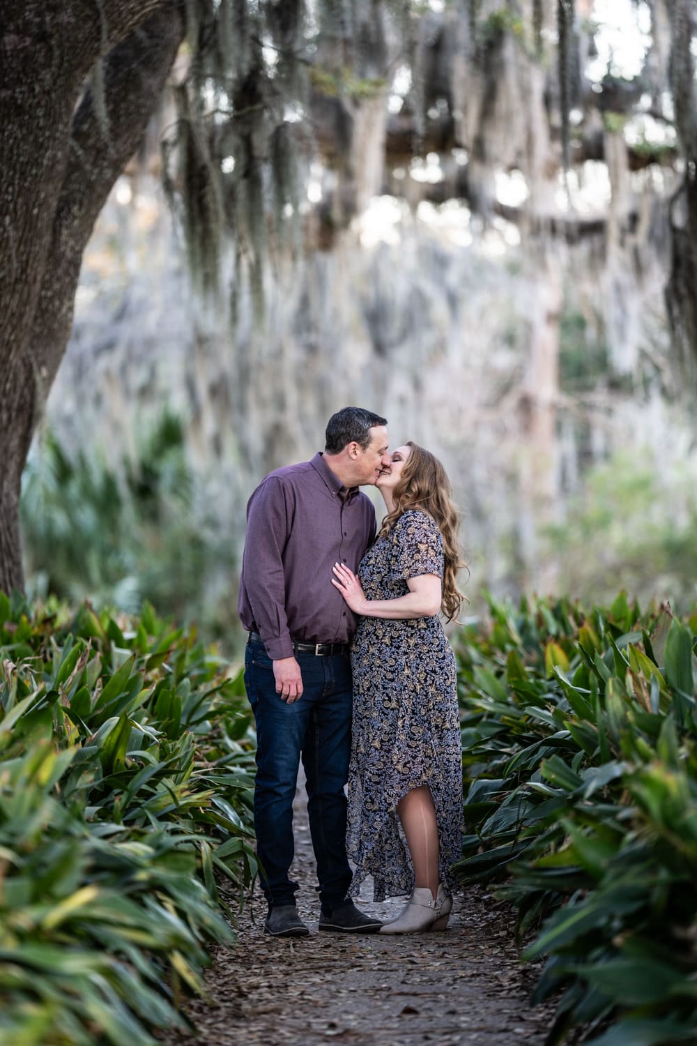 Couple kisses on walkway with moss-covered trees behind them