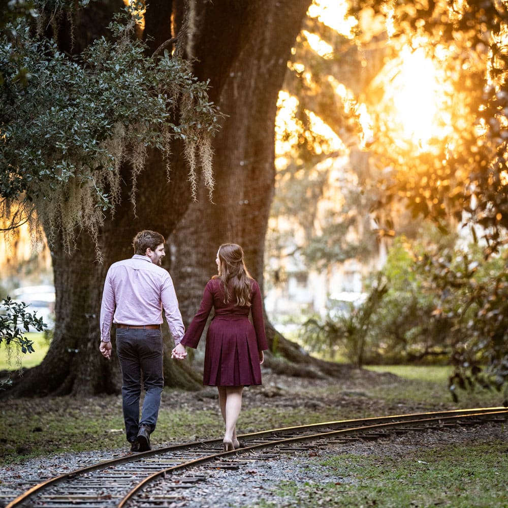 Engagement portrait of couple walking toward sunset along railroad tracks