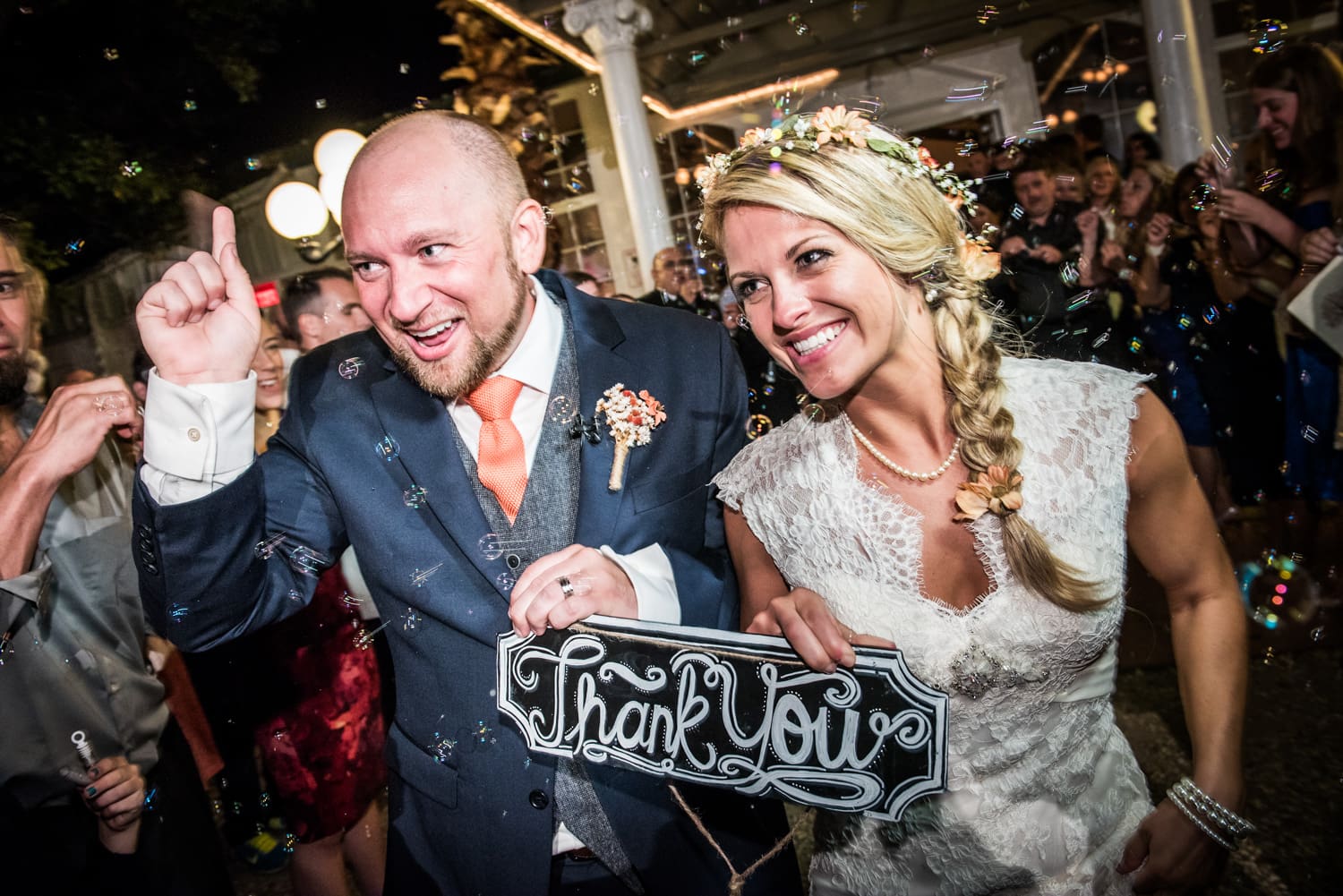 Couple departing wedding through bubbles, holding thank you sign