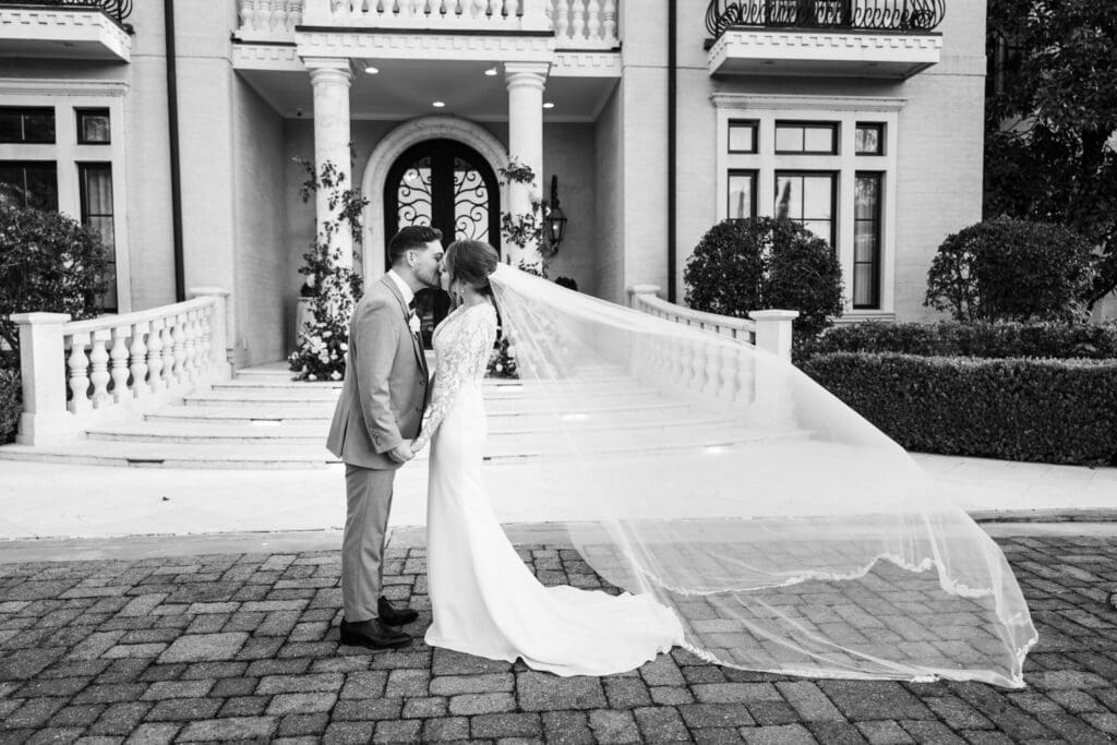 Bride and groom kissing in front of large home as veil blows in the wind