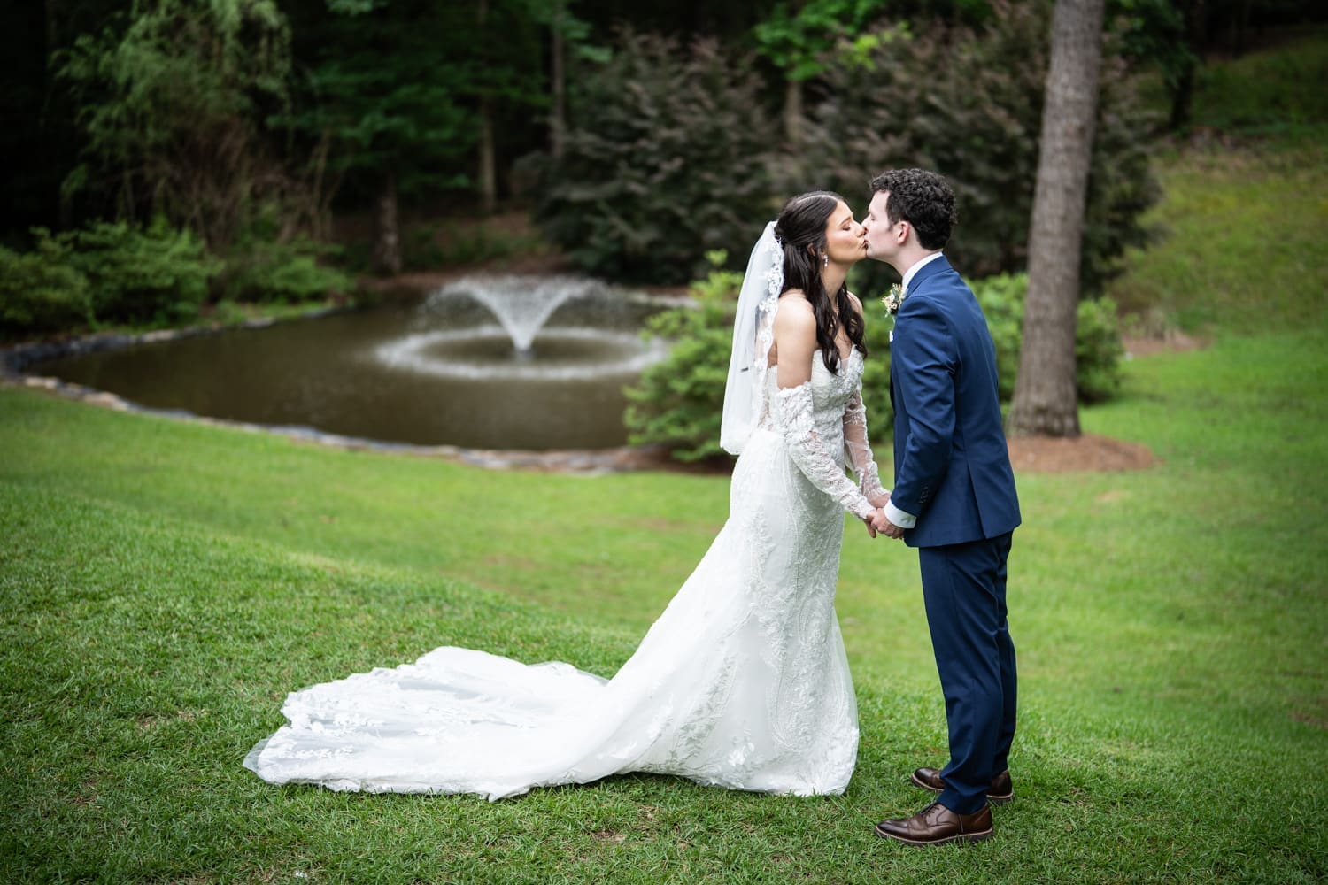 Bride and groom kissing on hilly lawn at Dogwood Venue with pond and fountain in the background